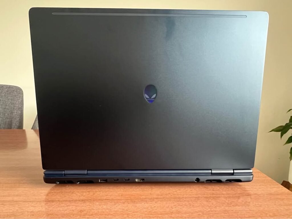 Rear view of the Alienware Aurora 16 Intel Core 7 gaming laptop with lid closed, showcasing the illuminated blue alien head logo on the dark gunmetal chassis, along with the rear I/O panel featuring USB Type-A, USB Type-C, HDMI port, audio jack, and dual exhaust ventilation vents