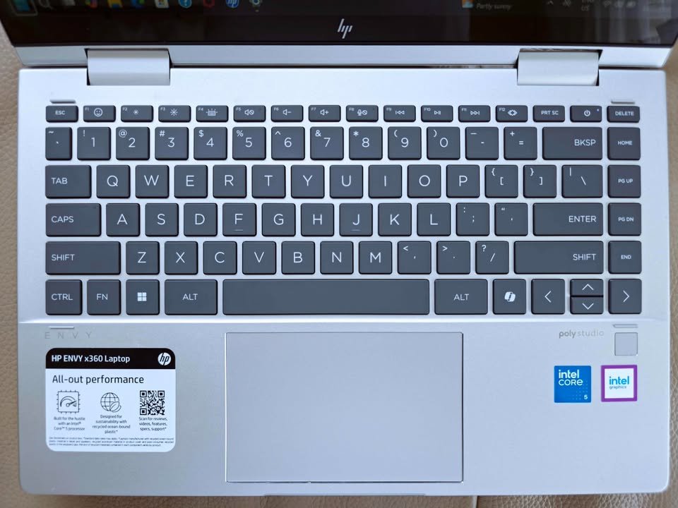 HP ENVY x360 2-in-1 laptop top view showing full-size dark gray keyboard layout with dedicated function row, large precision trackpad, Intel Core 5 processor badge, and HP ENVY x360 all-out performance product label